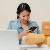 Woman checking package with phone near laptop and boxes